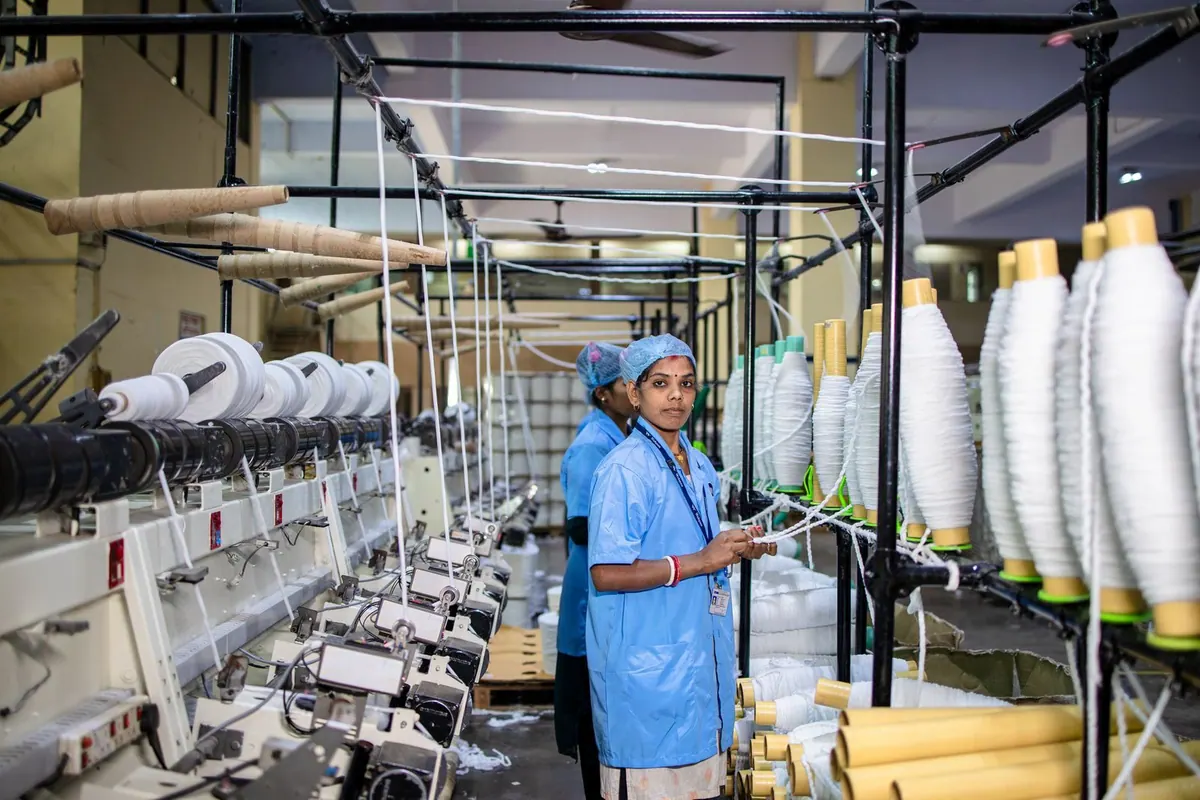 Mulher operando máquinas em uma fábrica têxtil, com carretéis de linha.