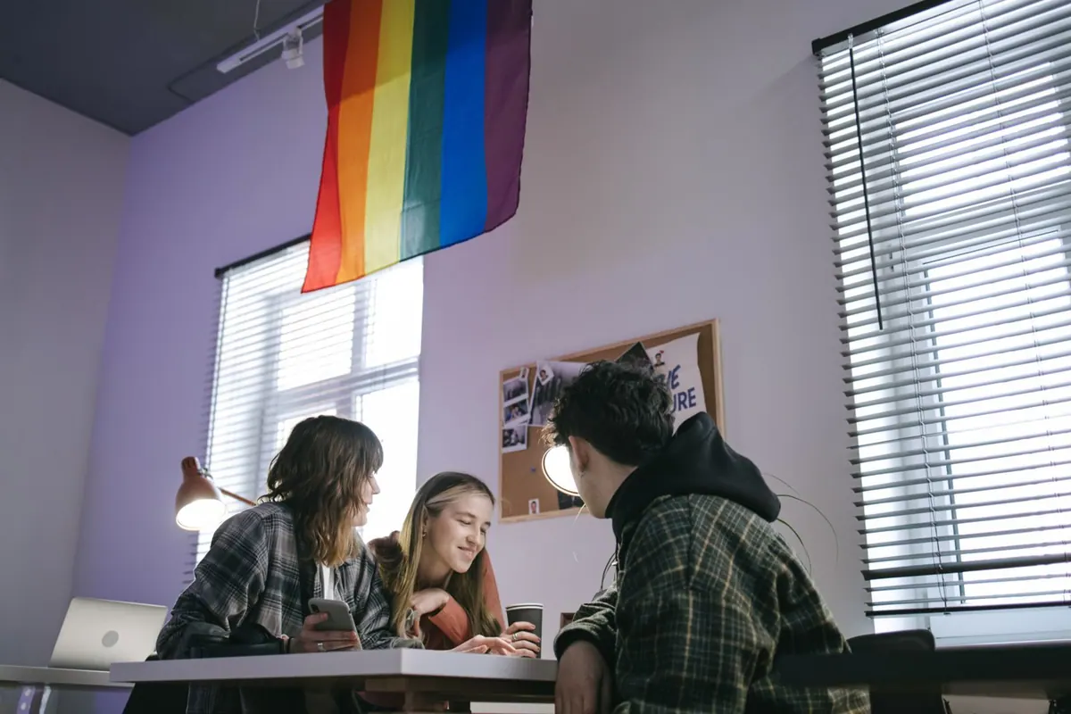 Três pessoas conversando em um escritório com uma bandeira LGBT+ pendurada.