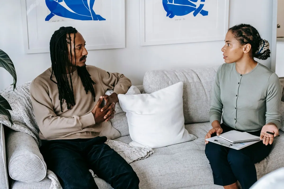 Duas pessoas negras sentadas em um sofá, conversando. Um homem com dreadlocks e uma mulher com um caderno.
