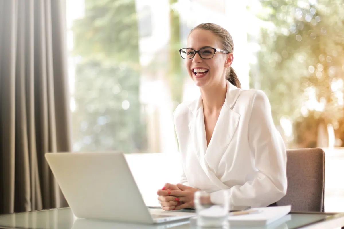 Mulher sorrindo com óculos sentada em frente a um notebook.