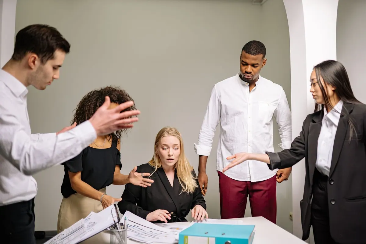 Colegas de trabalho expressam descontentamento em reunião, enquanto uma colega parece sobrecarregada.