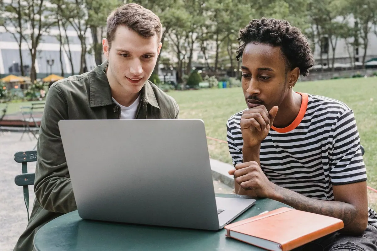 Dois homens diversos trabalhando concentrados em um laptop ao ar livre, em um parque.