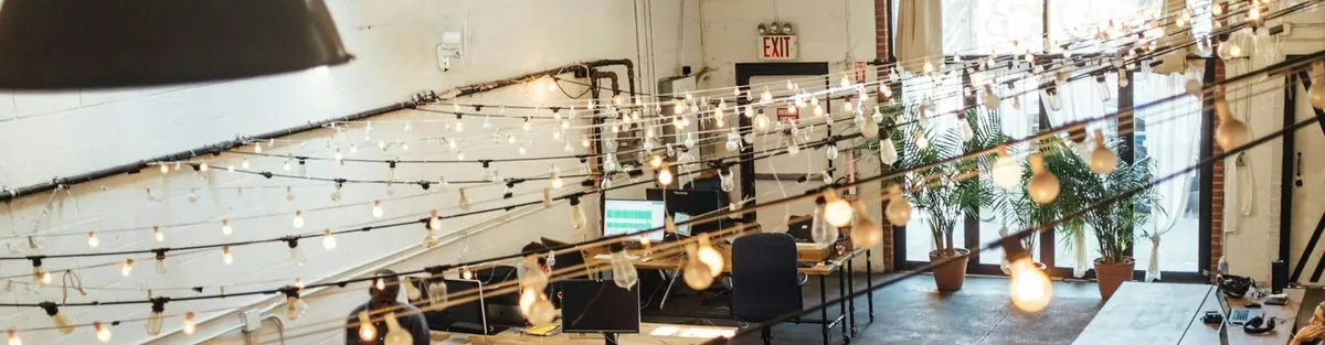 Escritório moderno com luzes penduradas, mesas e cadeiras, plantas e uma placa de saída.