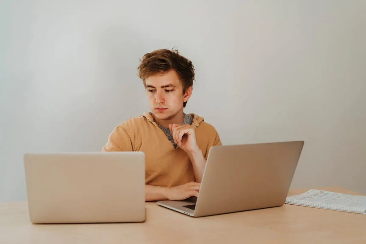 Homem jovem trabalhando em dois laptops simultaneamente em uma mesa de madeira.