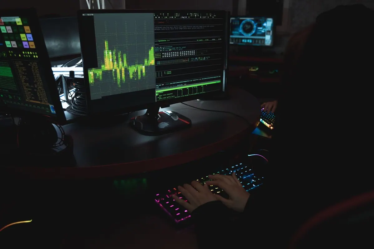 Pessoa digitando em teclado iluminado, com monitores exibindo gráficos e códigos.