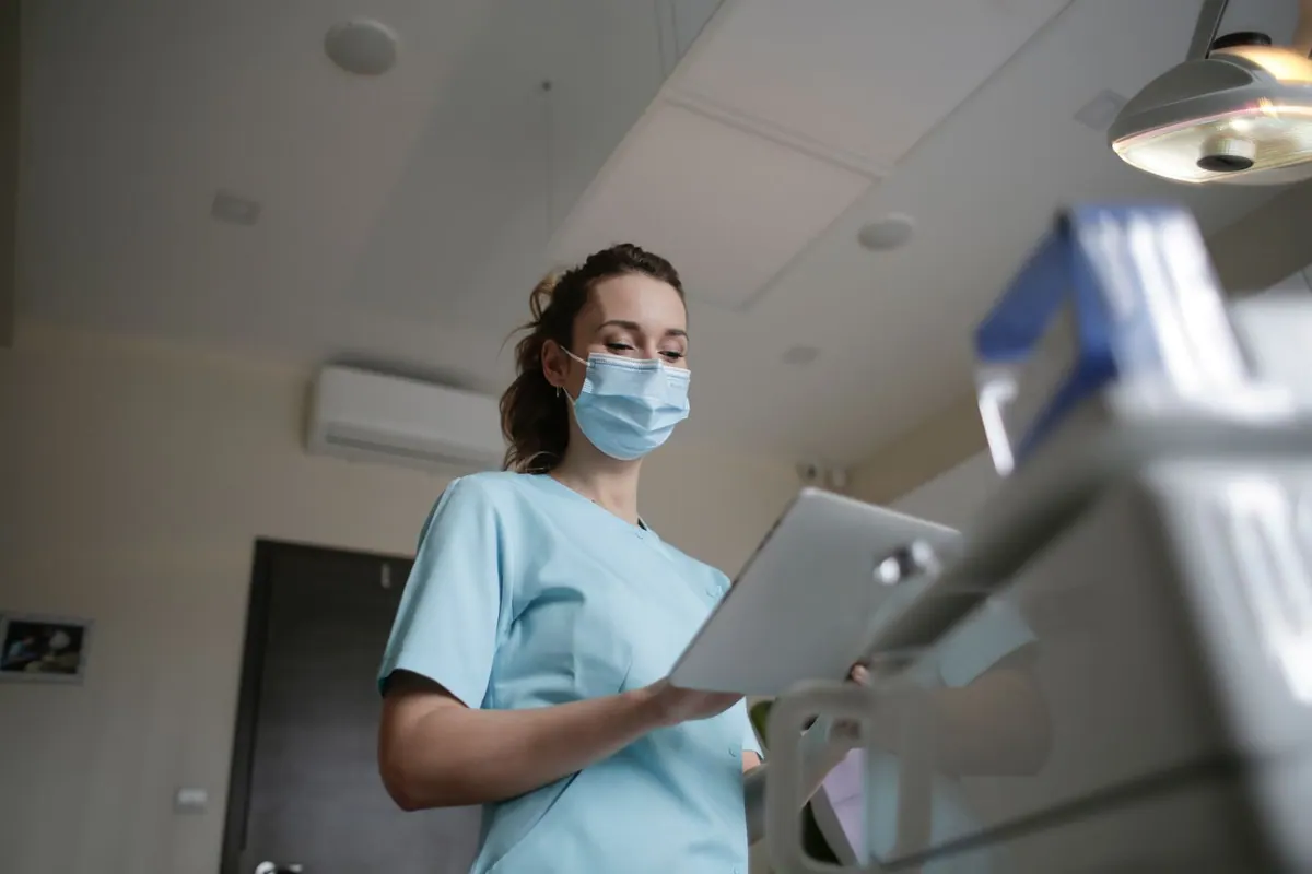 Profissional de saúde com máscara e uniforme, segurando uma prancheta em ambiente médico.