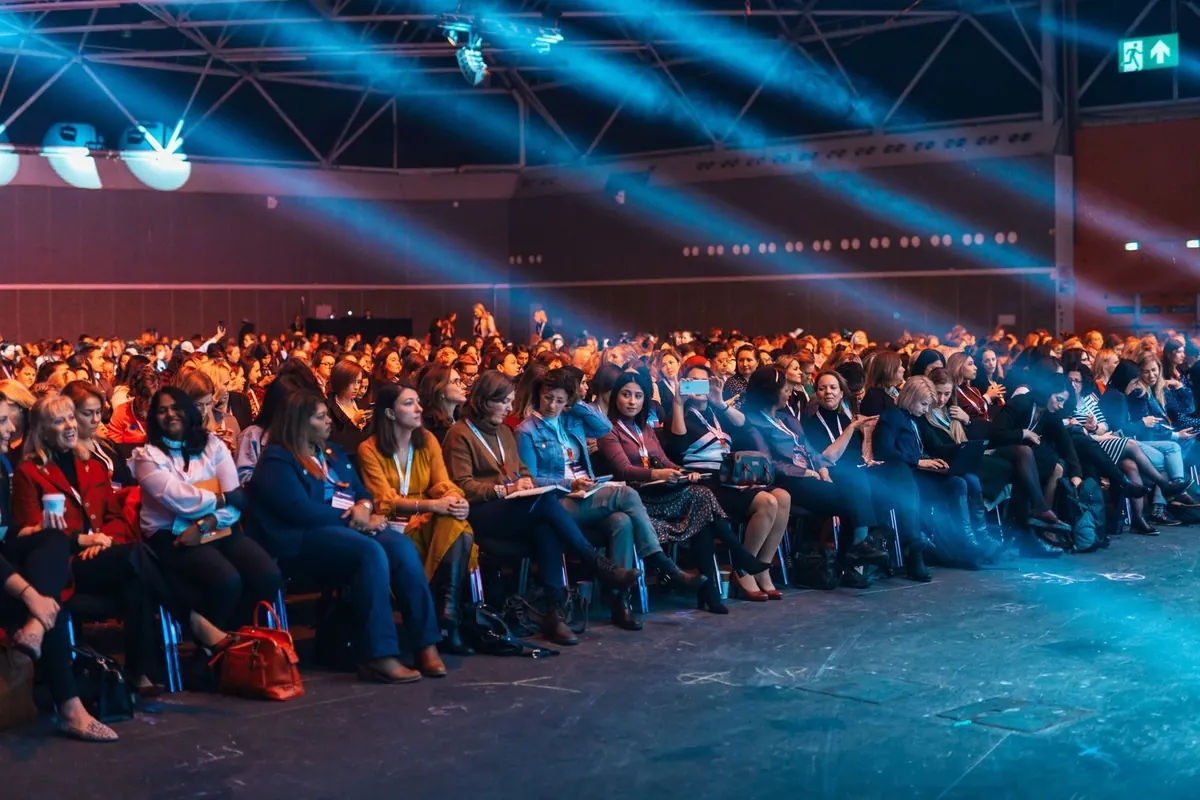 Pessoas sentadas em cadeiras em um evento, com iluminação azul no palco.