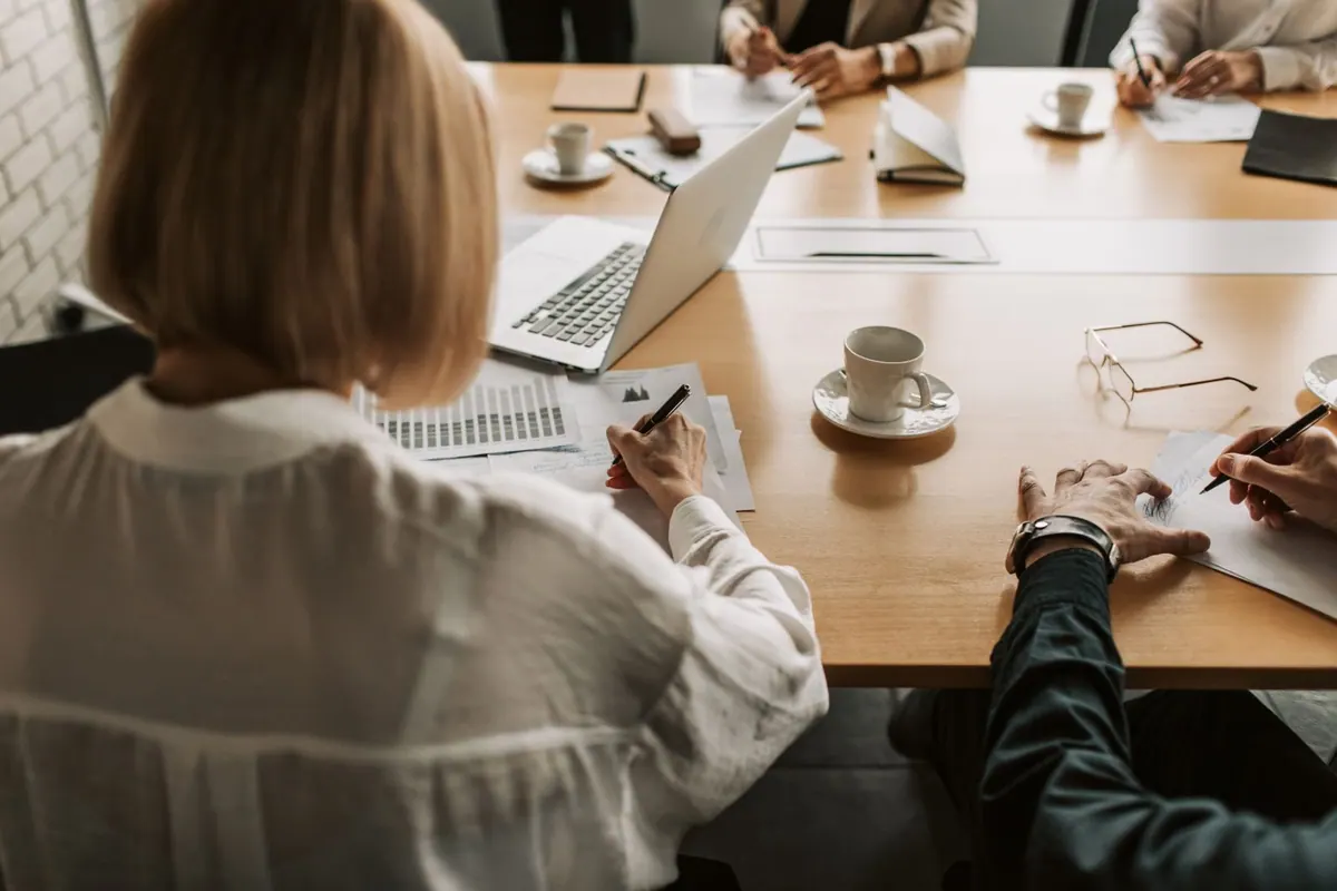 Pessoas em uma mesa de reunião, com laptops e documentos, avaliando projetos.