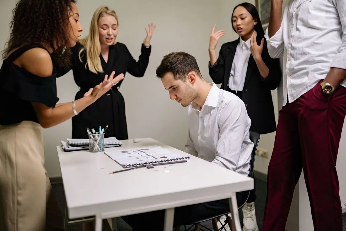 Pessoas discutindo com um homem sentado à mesa, que parece chateado.