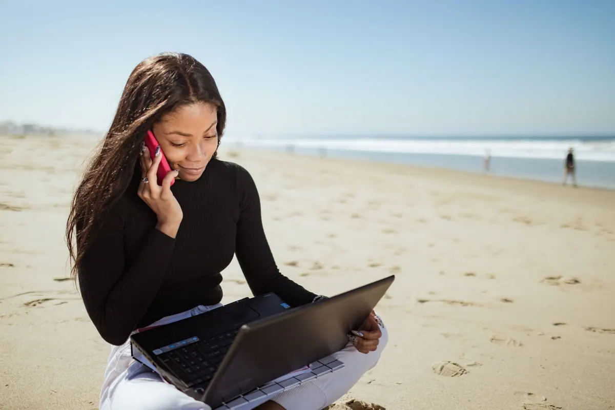 Mulher na praia, ao telefone e com laptop, simbolizando dificuldade em desconectar.