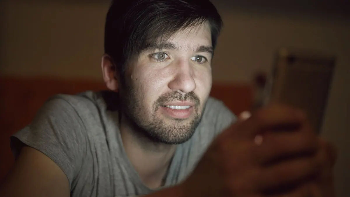 Jovem adulto com barba olhando fixamente para um smartphone à noite.