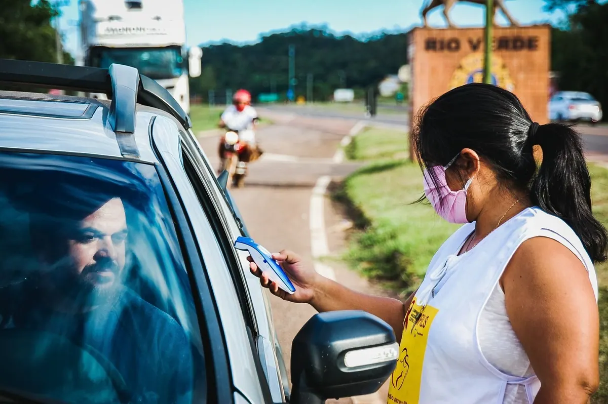 Mulher com máscara mede temperatura de motorista, refletindo impacto da pandemia.
