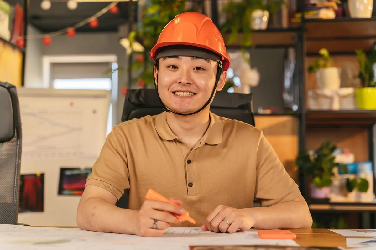 Homem asiático sorrindo, de capacete, sentado à mesa com caneta marca-texto.