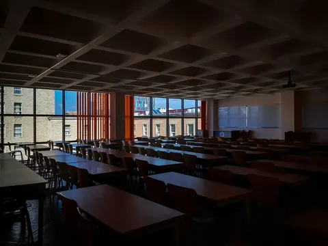 Sala de aula vazia com mesas e cadeiras, janelas grandes e quadros brancos.