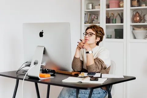 Mulher de óculos sentada à mesa do escritório, olhando pensativamente para a tela do computador.