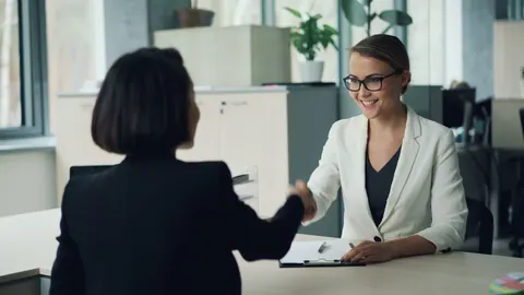Duas mulheres de negócios sorridentes apertando as mãos em um escritório moderno e profissional.