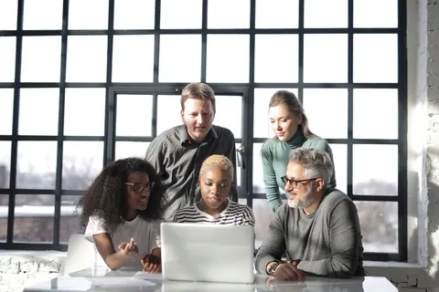Grupo diverso de pessoas colaborando em torno de um laptop em um escritório moderno.
