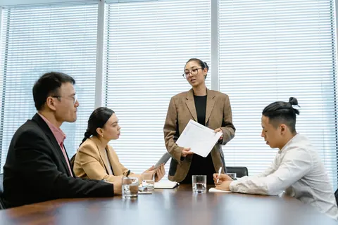 Mulher em pé apresentando documentos a colegas sentados em reunião de negócios.