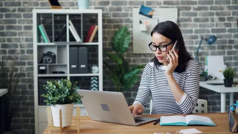 Mulher trabalhando em casa, usando laptop e telefone, com expressão séria.