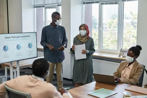 Pessoas em reunião de escritório com máscaras, discutindo gráficos em uma tela.