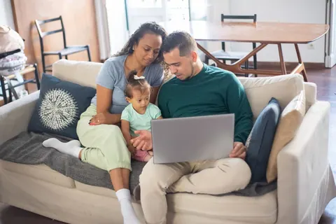 Casal com bebê no colo sentado no sofá, olhando para um laptop.
