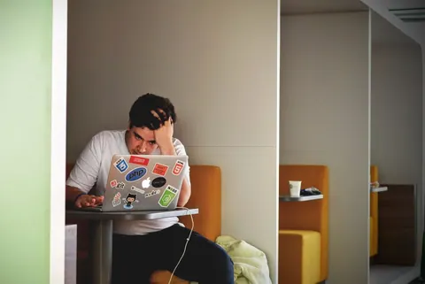Homem com expressão de frustração, mão na cabeça, olhando para um laptop cheio de adesivos.