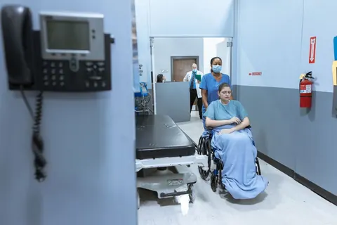 Enfermeira empurra paciente em cadeira de rodas em corredor de UPA.