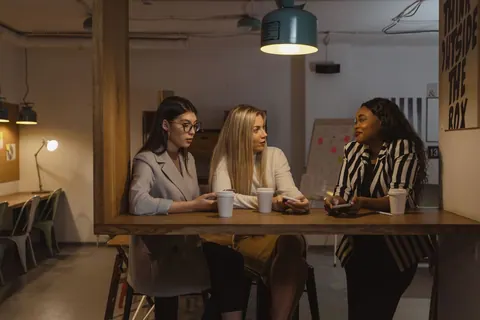 Três mulheres conversando animadamente em um ambiente de trabalho descontraído.