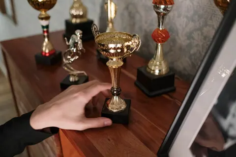Mão de pessoa tocando um troféu dourado em uma estante com outros prêmios.