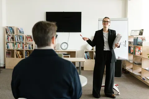 Mulher ensinando em sala de aula, com um aluno na frente.