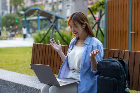 Mulher jovem sorrindo em videochamada no notebook, trabalhando remotamente em um parque.