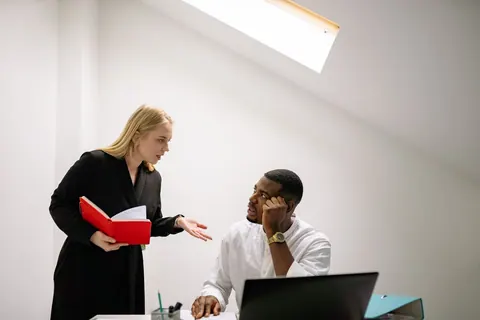 Mulher loira gesticula enquanto fala com homem negro em escritório.
