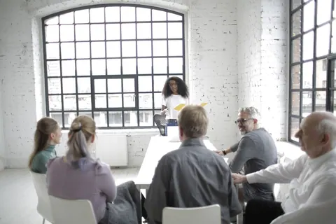 Uma mulher negra de pé, apresentando para um grupo de cinco pessoas sentadas em uma sala iluminada.