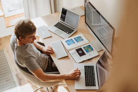 Jovem de camiseta cinza trabalhando em uma mesa com vários computadores e cadernos.
