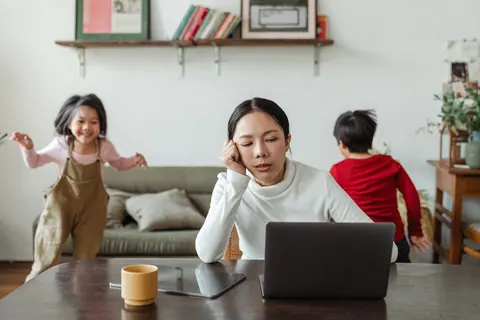 Uma mulher exausta trabalhando em um laptop enquanto duas crianças brincam ao fundo.