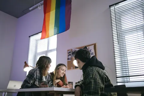 Três pessoas conversando em um escritório com uma bandeira LGBT+ pendurada.