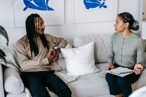 Duas pessoas negras sentadas em um sofá, conversando. Um homem com dreadlocks e uma mulher com um caderno.