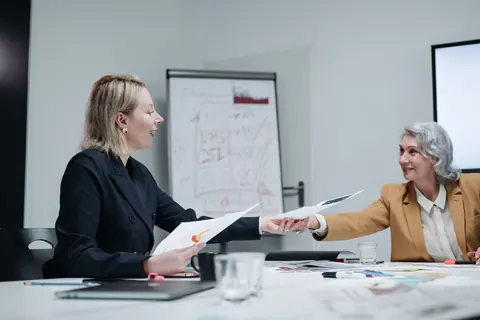Duas mulheres em uma reunião, uma passando documentos para a outra.