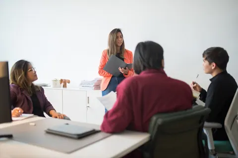Mulher em pé lidera reunião com três colegas sentados em escritório moderno.