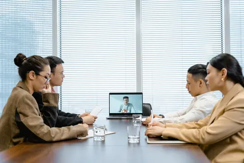 Pessoas em uma sala de reunião com um colega participando por videochamada no laptop.