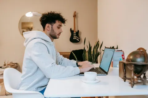 Homem jovem de moletom cinza trabalhando em seu laptop em casa.