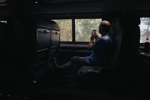 Homem idoso sentado em trem, olhando pela janela e usando celular.