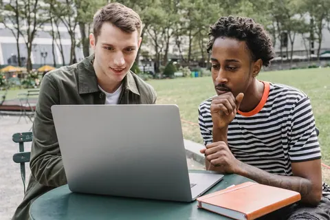 Dois homens diversos trabalhando concentrados em um laptop ao ar livre, em um parque.