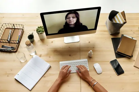 Mãos digitando em teclado branco, com monitor exibindo videochamada de mulher, em mesa de madeira.