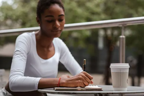 Mulher de pele escura escrevendo em um caderno com uma caneta, com um copo de café ao lado.