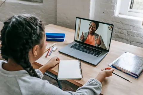 Uma pessoa com tranças assiste a uma videochamada em um laptop, com cadernos e lápis.