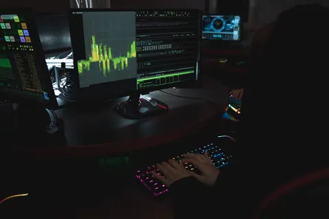 Pessoa digitando em teclado iluminado, com monitores exibindo gráficos e códigos.