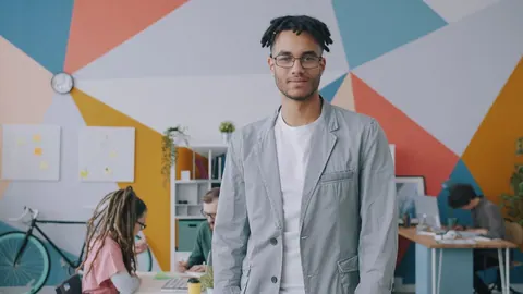 Homem sorridente em escritório moderno com colegas trabalhando ao fundo.