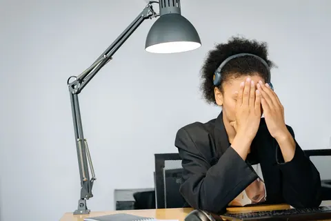 Mulher com fones de ouvido cobrindo o rosto com as mãos, expressando exaustão.