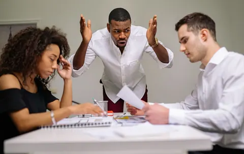 Chefe bravo gesticula para funcionários em uma mesa de escritório.